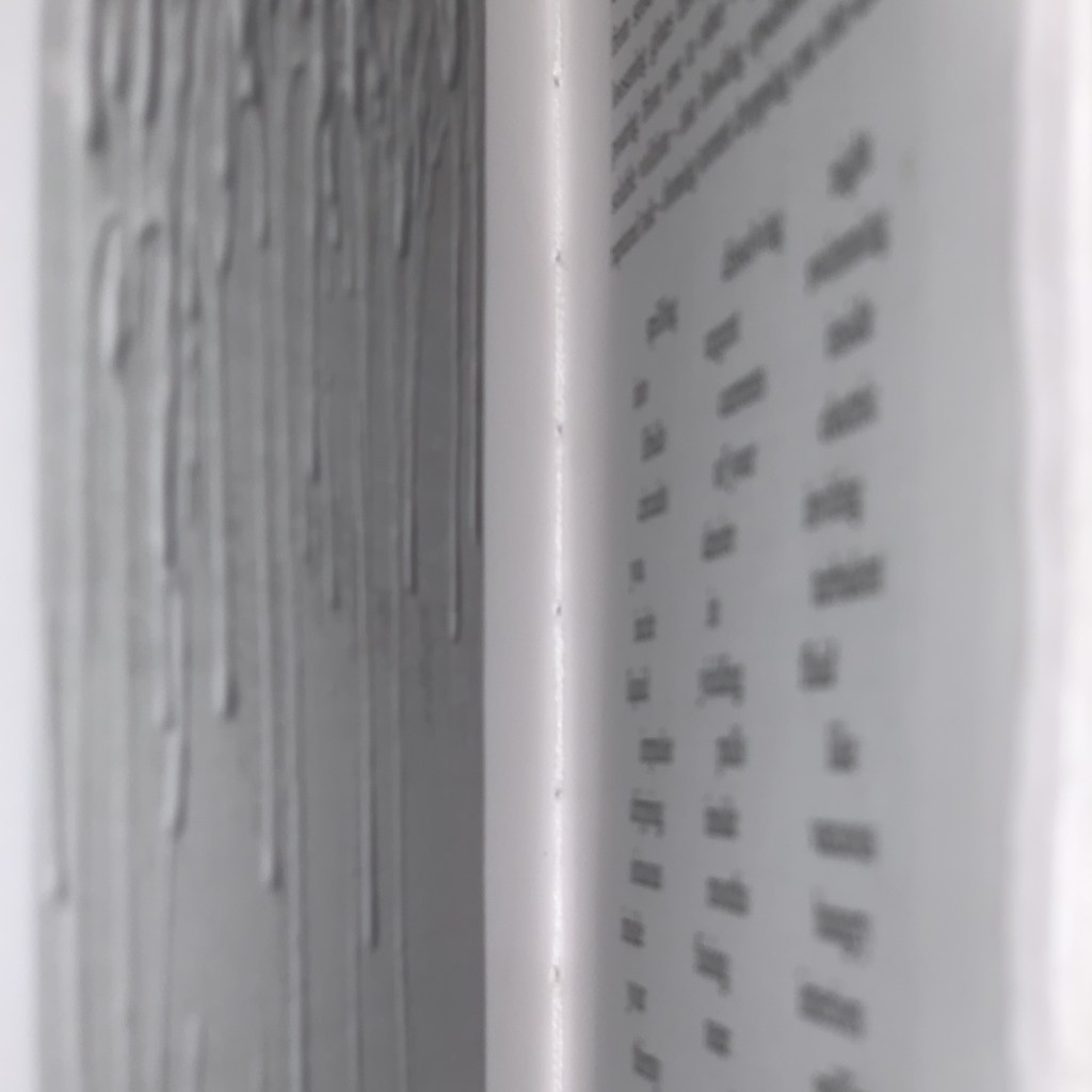 Out of focus image of two pages in a book, on the left an image of textural white drips, on the right a poem with three waving columns.