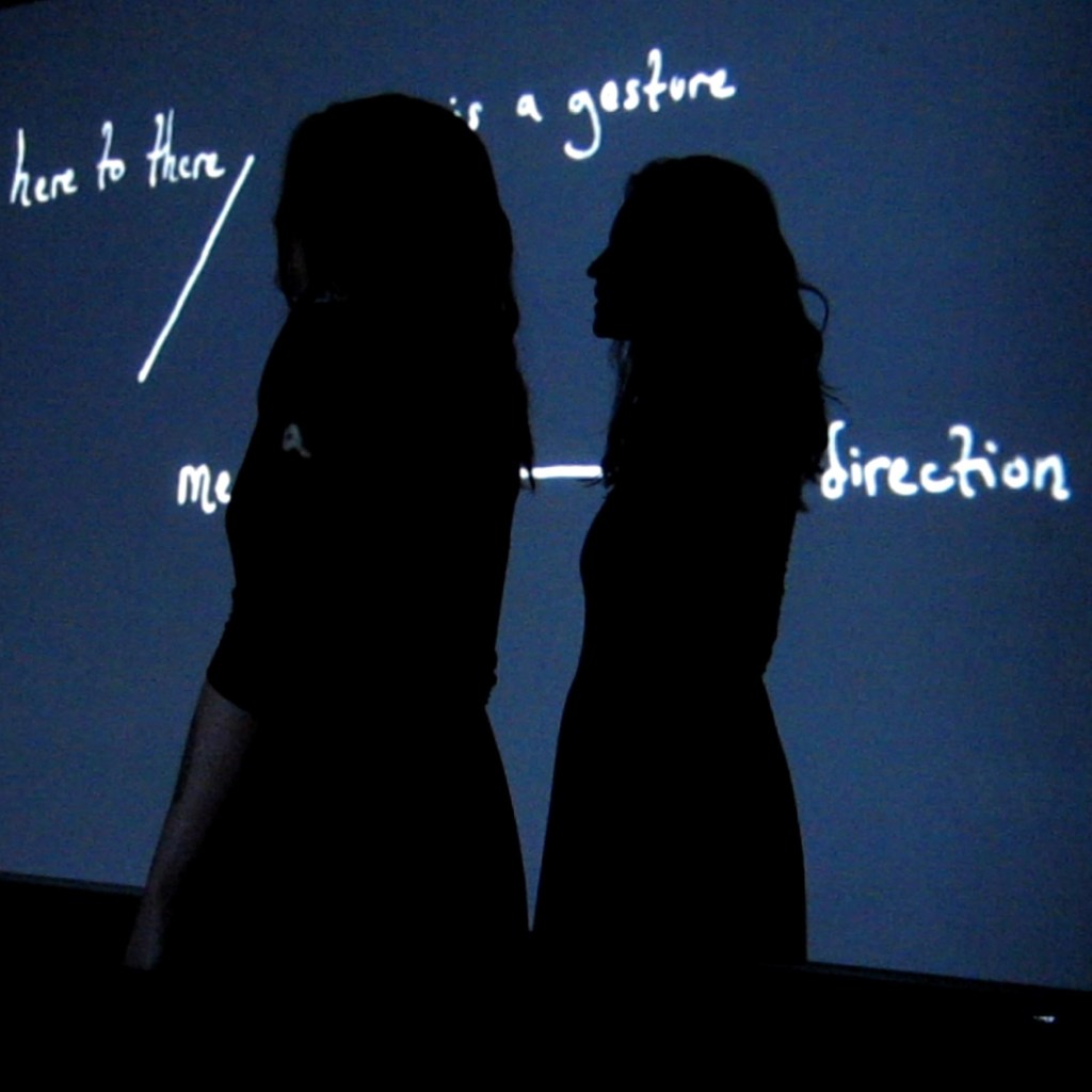 The doubled silhouette of a person against a projected image of writing arranged in a diagram, which is partially legible behind the figure, reading "here to there / is a gesture / me - direction"